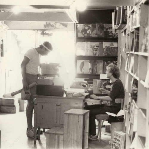 Intérieur de la Librairie L’Androgyne. Les murs sont tapissés de livres. On aperçoit une personne à gauche feuilleter un livre, une personne regardant quelque chose au bureau à l'entrée, et une troisième personne assise derrère la caisse. Fonds Librairie L’Androgyne/Androgyny Bookstore. Collection des Archives gaies du Québec.